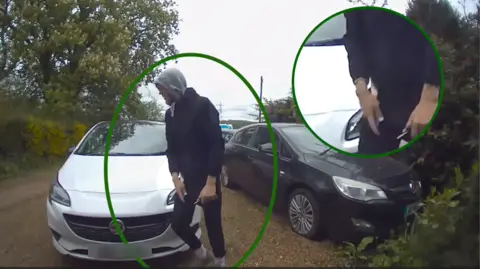 Hertfordshire Police A man wearing a black top and bottoms with a grey hood holding a knife while walking in front of a a silver car and a black car