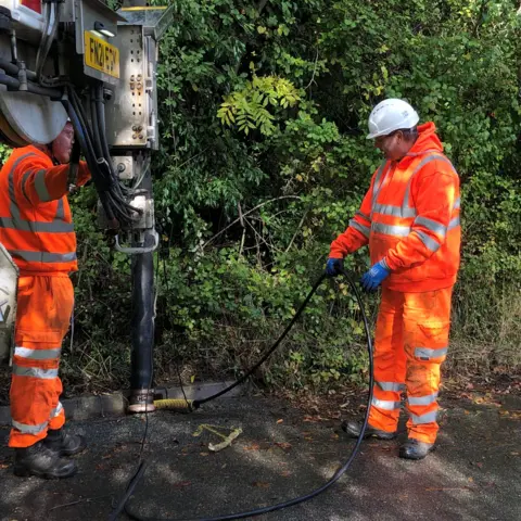 Vikki Irwin/BBC Two men in orange protective suits and hard hats clear out a roadside drain using specialist equipment. The path borders extensive green-leaved bushes. 