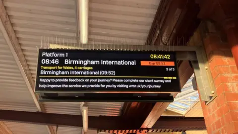 A virtual board attached to a brick wall at a train station. On the display is information for a train to Birmingham, by Transport for Wales. It says it is delayed by 11 minutes.