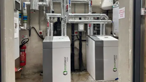 Machines in the plant room at Randalstown apartment block. They are white and grey cubes with green and grey writing saying "Ecoforest" printed on them vertically - along with a tree logo. Silver pipes are protruding from them.