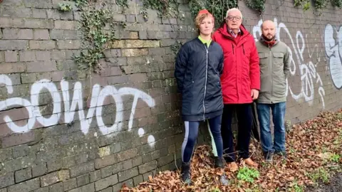 Three people wearing khaki green, red and blue raincoats stand by a wall covered in graffiti with a solemn expression on their faces