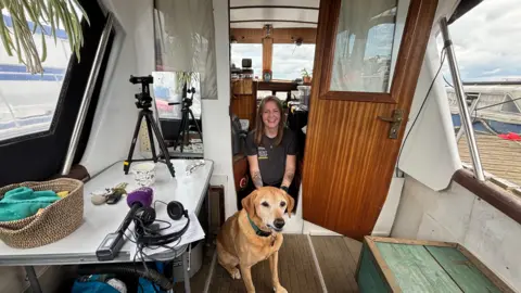 Luke Deal/BBC Holly Brookes crouches in the doorway into her houseboat. A golden Labrador dog sits in front of her.