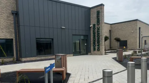 A grey cladded and light stone building sits on a square centre. The side of the building reads in green 'Workington Diagnostic Centre'.