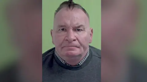 IOM Police A police mugshot of Mark Thone, a man with receding grey and black hair. He is wearing a dark grey jumper with a checked shirt underneath. His mouth is turned down at the corners and one eyebrow is raised as he looks directly at the camera.