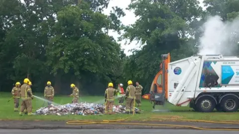 Breckland Council Firefrighters in khaki uniform and yellow helmets use hoses on a mound of rubbish spread across grass. Parked side-on to their right is a white bin lorry billowing smoke