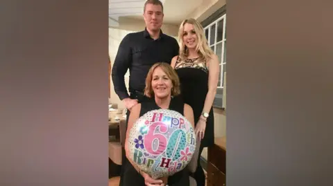 Family photo Jane Blackwell holds a 60th birthday balloon and is sitting down. Her daughter, who is wearing a black dress, and son, who is wearing a black shirt, stand behind her. 