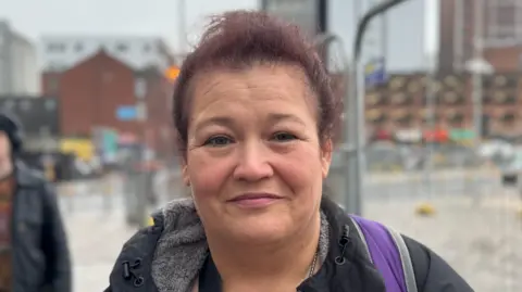 Margie is smiling at the camera. She has purple hair and is wearing a black coat and purple backpack. She is standing outside a train station in Belfast.