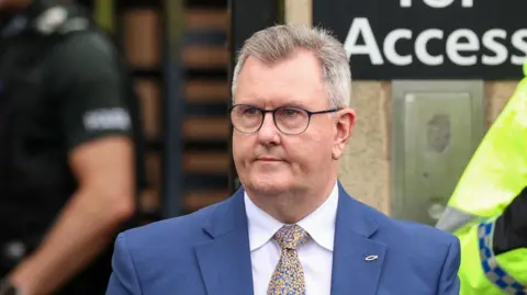 Reuters Jeffrey Donaldson. He has short grey hair, wearing glasses, a blue blazer, white shirt, blue and yellow tie, with a fish pin on his lapel.
