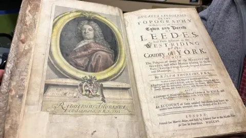 An historical open book on display. On the left page is a coloured portrait of a man with long, curled hair, dressed in early 18th‑century clothing. Printed text identifies him as  Radulphus Thoresby, Leodiensis, S.R.S. 1712. The right page shows the book’s title page, filled with  ornate text. The title reads 'Ducatus Leodiensis'.