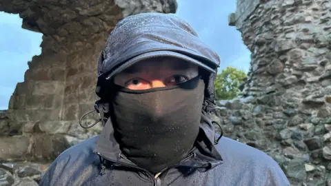 A man looking directly into the camera. He's wearing a balaclava and a cap so you can only see his eyes. In the background you can see castle ruins - the photo is taken in the evening so the sky is starting to darken. 