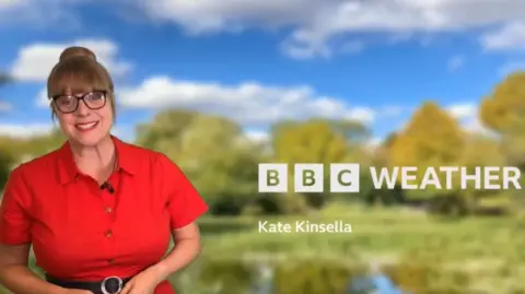 Weather presenter Kate Kinsella wearing a bright red dress and black belt, standing in front of the BBC TV weather backdrop with blue skies and green countryside