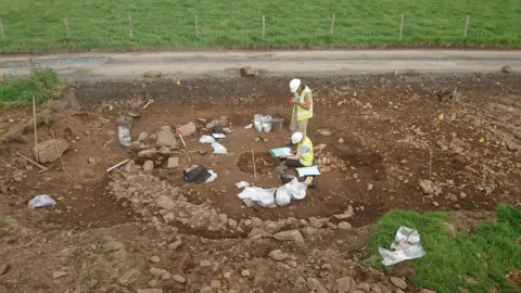 Guard Archaeology Two people in white hard hats and yellow high visibility tops look into a hole in the earth surrounded by tools with a fenced green field in the background