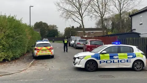 BBC A street with cars parked on both sides of the road. Two police cars and an officer are in the image.