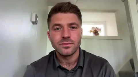 A man with short brown hair and stubble - he wears a grey shirt and sits next to a staircase with a window behind him in a screengrab from a videocall.