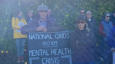 Ella Harget-Dash/BBC Paul and Jackie Thompson are facing the camera with expressionless faces. They are standing with a black sign that reads, in white writing, 'National Grids 180km mental health crisis'. Other protesters can be seen in the background.