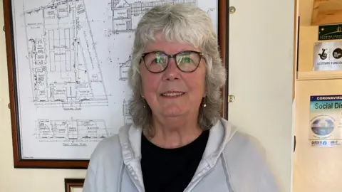 A woman with shoulder-length grey hair and glasses, smiling.