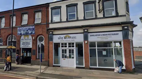 A view of a takeaway called Ali's Kitchen on a street with a bus stop. The restaurant has a white door and signage. 