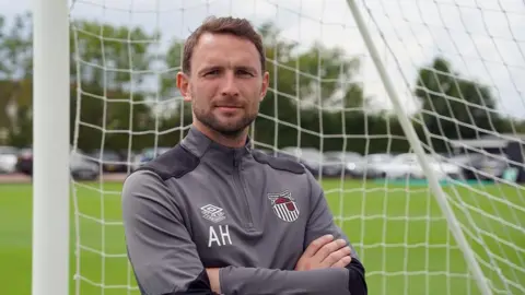 Grimsby Town FC Alex Hughes posing for a picture while stood in a goal wearing a Grimsby Town tracksuit.