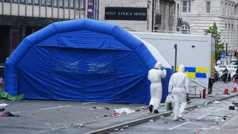 PA Media Two forensic investigators in white full-body overalls walk away from the camera along a road next to a large inflatable blue police tent. A black sign on a building in the background reads 'West Africa House'.