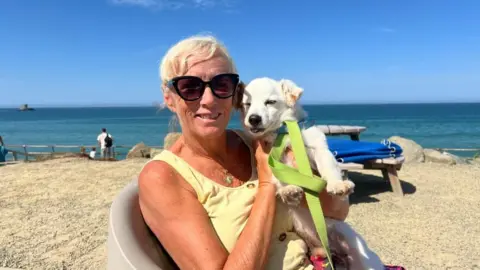 A woman with short blonde hair holds her small white dog to pose for the camera. She's wearing large black sunglasses, and a yellow, sleeveless top. The dog has a blue collar and a green lead. They're sitting on a chair in a gravel area with the sea in the background.
