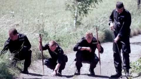PA Media Four police officers in a line, looking for evidence on a country lane. They are dressed in black police overalls and two are carrying sticks. Three are crouched down looking at the ground, while one is standing with what appears to be a metal detector.