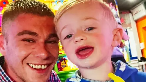 Nik Russell rests his forehead on his son's head as they both smile at the camera. Nik has short cropped brown hair and Leon has short blonde hair. They appear to be in a brightly coloured arcade.