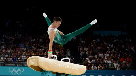 Reuters McClenaghan on a pommel horse, mid movement holding himself up by one arm. The horse has the Paris 2024 logo on it, and behind Rhys is a stand full of people. 