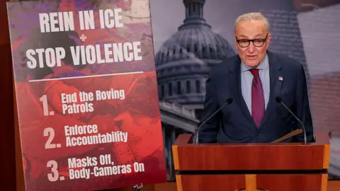 Getty Images Senate Minority Leader Chuck Schumer speaks at a press conference standing next to a board with list of three demands from Democrats.