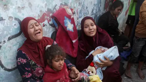 Reuters Two women cry out as they lean against a wall and hold two children - one a young girl and the other a baby wrapped in a blanket - in a school sheltering displaced people in Gaza. Another young woman is seen in the background.