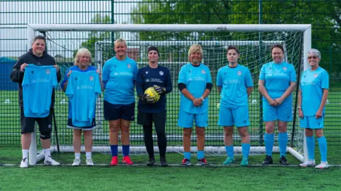 Simon Thurlow Woman's walking football team stood holding blue shirts in front of a goal on a football pitch. There are trees in the background.