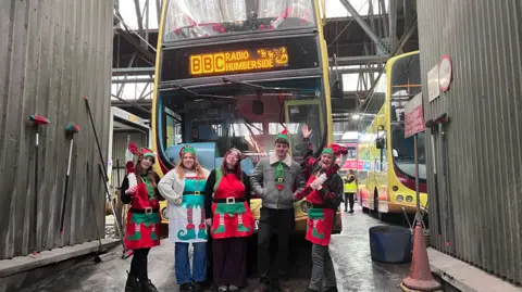 Five people dressed in Christmas fancy dress stand in front of a double-decked bus at a bus depot - the bus's display reads 'BBC Radio Humberside' and has Santa with his sleigh. 