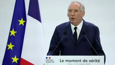 Francois Bayrou stands in front of a lectern reading "The moment of truth". Behind him are a French and an EU flag