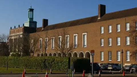 County Hall in Hertford - a red-brick building with arched ground-floor windows and a central tower featuring a cupola, seen on a clear day with a hedge in front of the building and cars parked to the right.