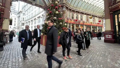 Leadenhall Market with people, including men in suits, walking past a Christmas tree on cobblestone streets. 