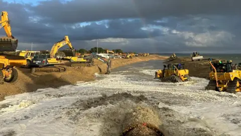 Southsea Coastal Scheme Yellow and black diggers and dumpers on a beach and in the sea water moving shingle with a pipe seen with shingle being pumped on to the beach area.