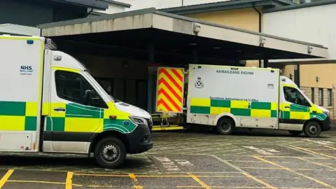 Ambulances with green and yellow checked markings, parked outside a hospital building.