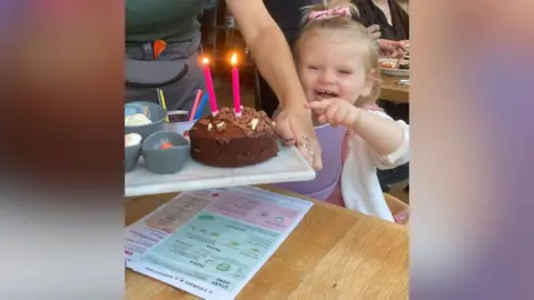 Sarah Normington A two-year-old girl is being given a chocolate birthday cake with two candles on it. She is sitting at a table in a restaurant with a menu on the table in front of her.