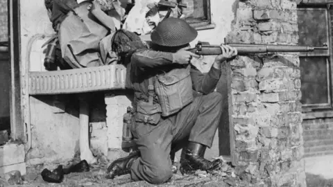 A black and white image of a soldier crouching down beside a wrecked building. He points a Lee Enfield rifle, his eye lining up the sights, finger on trigger.