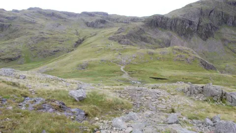 Looking back down on Sty Head while ascending Great Gable by the Breast Route. The path to Sprinkling Tarn is ahead centre. There is a rocky path leading up to a crag which is covered in green grass.