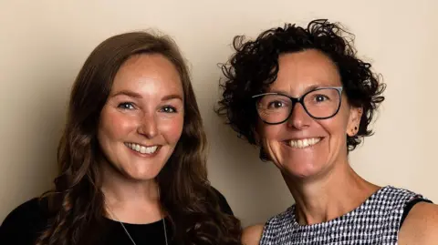 Two women. One with long dark, brown hair and one with short curly brown hair and glasses. They are both smiling at the camera.
