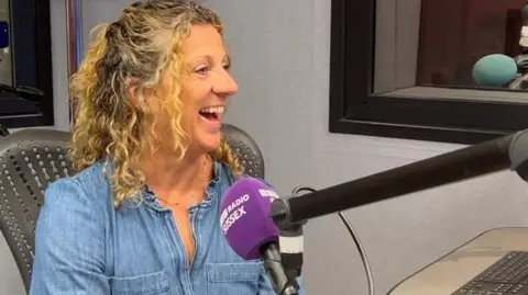 BBC Olympian Sally Gunnell, wearing a light blue denim shirt, sits in the BBC radio studios at Brighton, speaking into a microphone with a purple branded windsock cover.