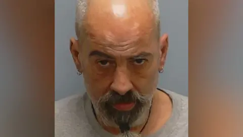 Kenneth Walker pictured in a mugshot. His head is looking down but his eyes are fixed on the camera. He has a bald head with short grey hair at the sides, ringed earrings in both ears, and a thick black and grey goatee beard tied at the bottom.