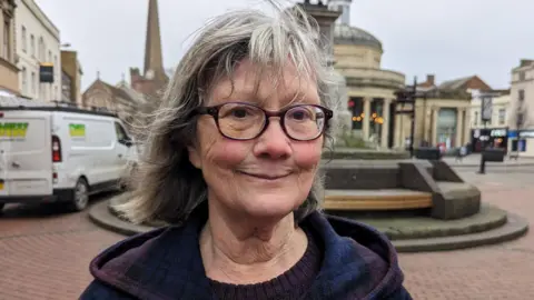 A woman with round glasses and a purple and blue coat stands in front of a statue in the centre of Bridgwater in Somerset