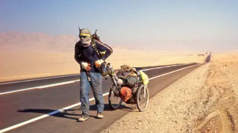 Karl Bushby A man with a shemagh wrapped tightly around his head, walks along a long, straight road in a desert. His clothes are dirty and he is pulling a two-wheeled trailer containing all his possessions.