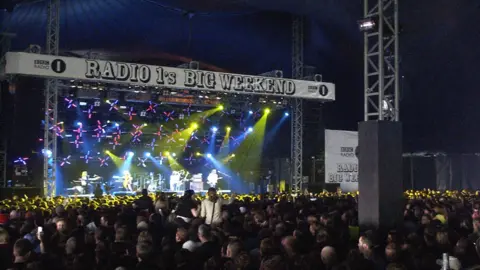 A large crowd of people inside a giant blue marquee looks at a stage, with Radio's Big Weekend branding around it.