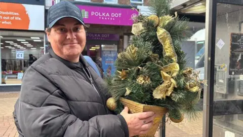 A woman wearing a black baseball cap, smiling and holding a mini Christmas tree dressed with gold ribbons and pine cones. She is wearing a black coat. Shops can be seen behind her.