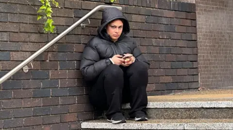 BBC News Fatimaalzhra Yousif, also known as Zara, sits on steps outside court wearing a black puffer jacket, black trousers and trainers. She is clutching a mobile phone and a vape
