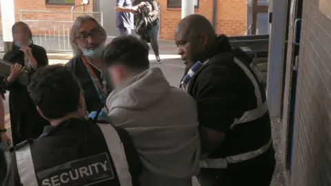 Channel 4 A television still, showing members of hospital staff, including Leston Scafe, holding a young man in a grey hoodie by the arms