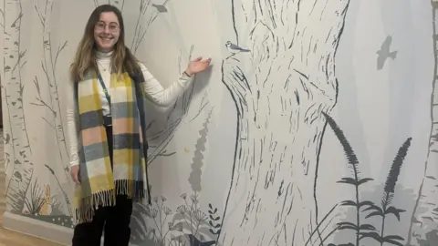 Severn Hospice A woman with long brown and blonde hair dressed in black trousers, a white top and a long navy, green, white and peach scarf around her neck. She is gesturing to a wall with a large mural on it. The mural shows trees, plants and flowers , with birds, rabbits and other insects and animals
