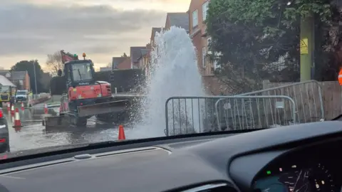 Andi-Emily Stokes A photo taken from the passenger side of a car showing water erupting from the road with an road digger and orange cones marking the area. 